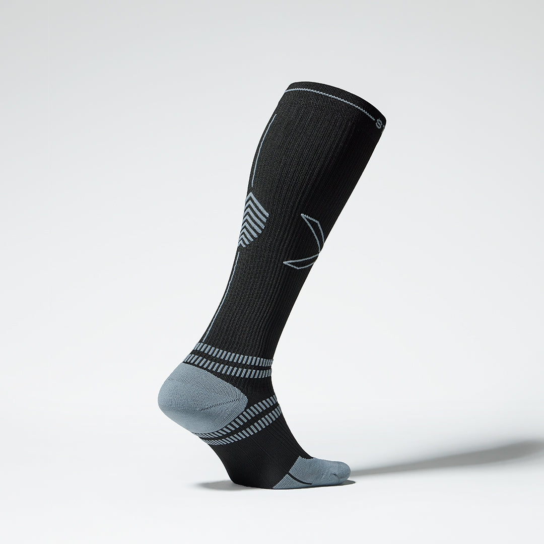 A standing black knee high compression sock with grey lines from the side.