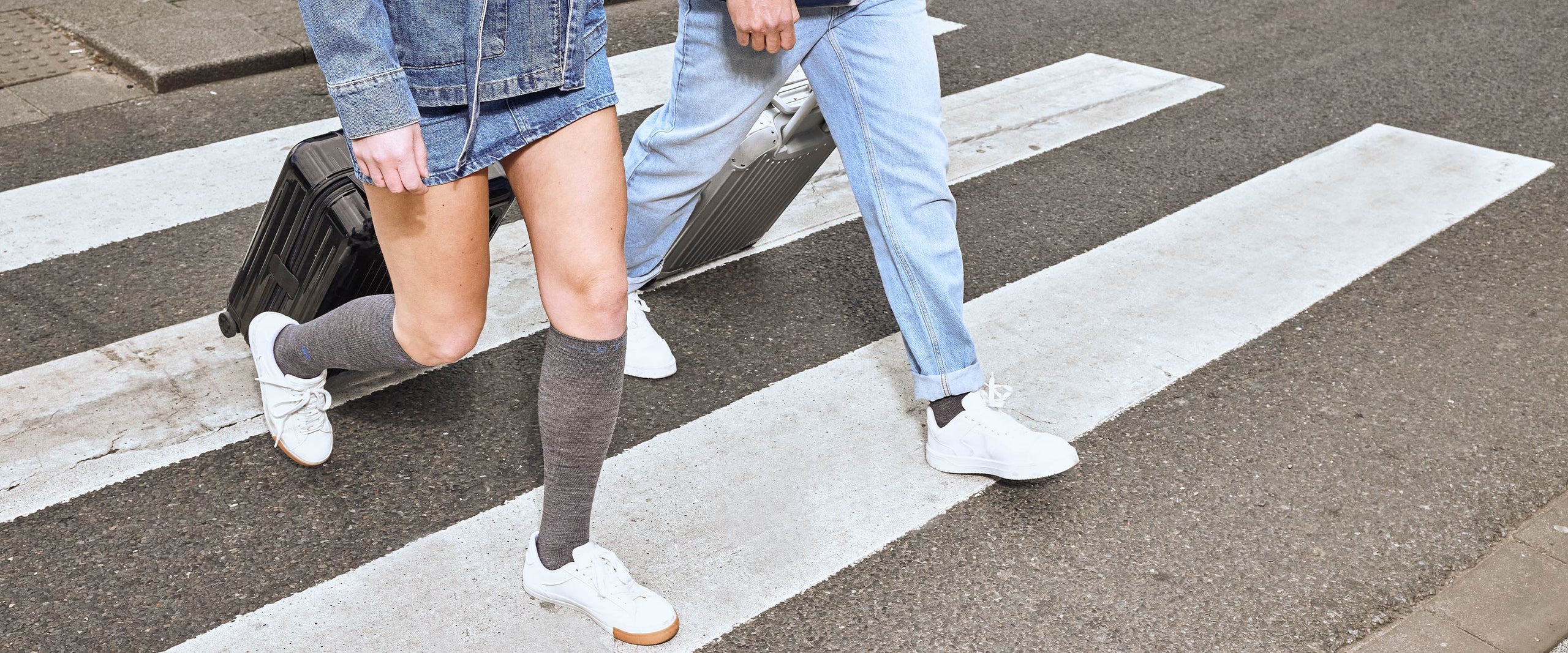Two people walking with suitcases wearing white sneakers and grey socks.