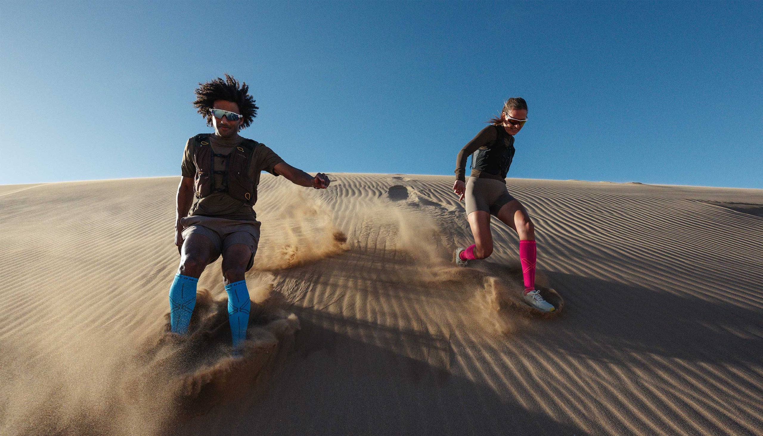 Two people running on sand dunes with a clear blue sky.