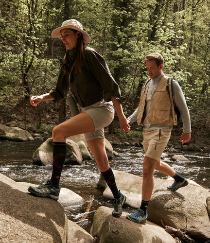 Man and woman hiking on rocks alongside riverbed.