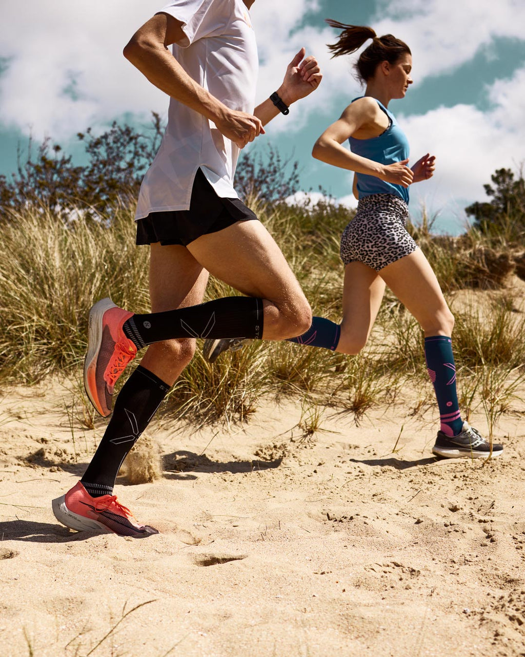 Legs of a man and woman running on sandy ground wearing a white and blue tank top