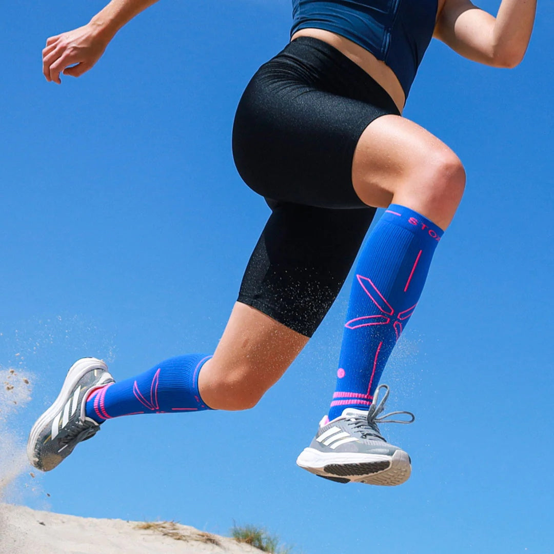 Legs of a woman running on the beach wearing blue knee high compression socks with pink details. 