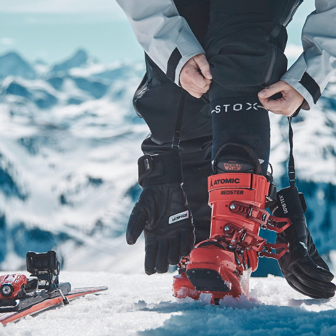 Person pulls up ski sock and wears red ski boots.