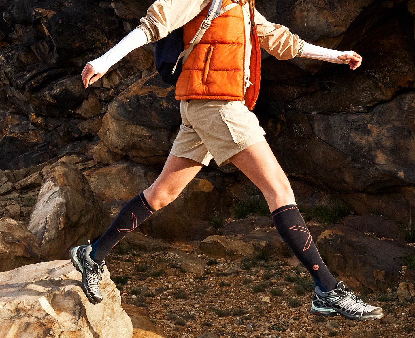 Woman jumping in the sun with dark blue compression socks and orange bodywarm