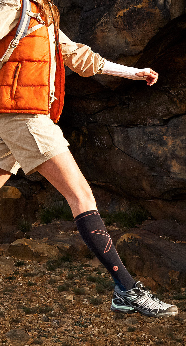 Detail shot of woman jumping in the sun with dark blue compression socks and orange bodywarmer.