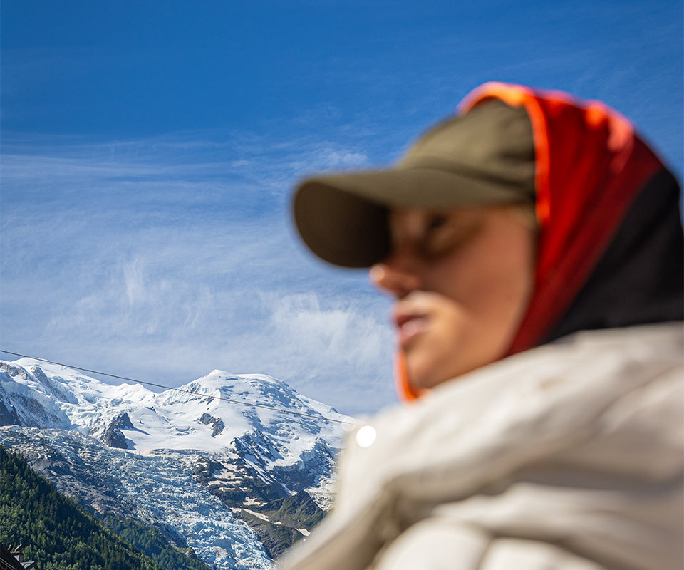 A woman wearing a cap and a red scarf around her head with mountains in the background.