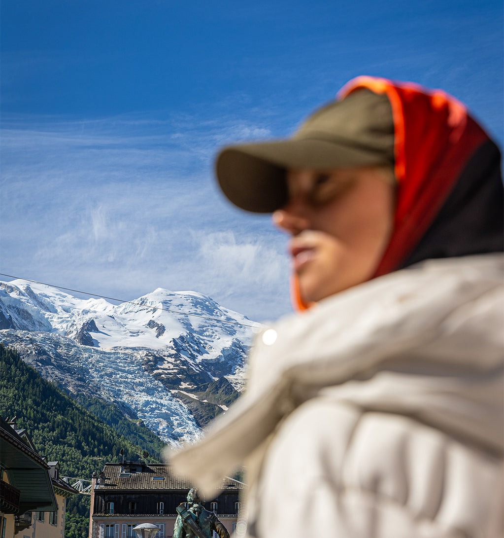 A woman wearing a cap and a red scarf around her head with mountains in the background.