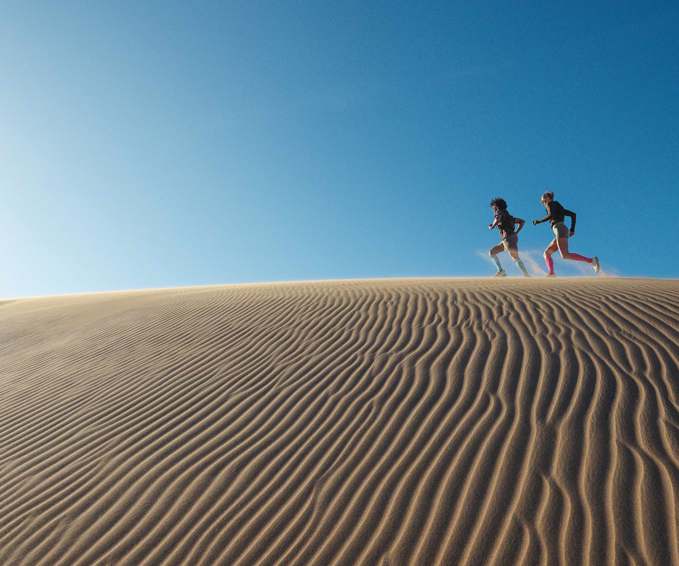 Two people running on a sand dune under a clear blue sky